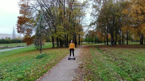 Woman ride hoverboard at city park, dolly shot, autumn season. She stay on self-balancing scooter and move along ground path, old green grass and maple trees on the way