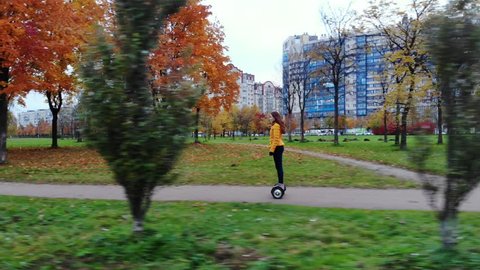SAINT PETERSBURG, RUSSIA - OCTOBER 12, 2018: Woman ride self balancing scooter at city park, side tracking shot. Coloured leaves on maple trees at autumn time, some buildings seen on background