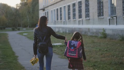 Happy mother with her daughter in school uniform go home. Mom and daughter are walking along the alley along the school. Slow Motion Sunset