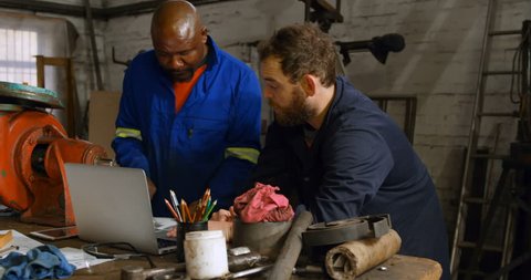 Two diverse  metalsmiths discussing over laptop on desk in workshop 4k