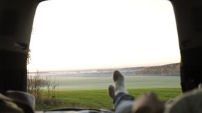 Young woman admiring the sunrise in trunk of car. Legs of girl dressed in woolen clothes against backdrop of forest panorama in fog . View from inside slow motion video