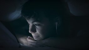 Boy watching movie or playing games on tablet computer at night. Child with headphones under blanket on bed using smartphone or tablet pc. Caucasian boy to make video call to talk to friends. 