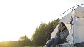 Camping young woman sitting in camp tent . Happy girl relaxing on vacation. Young female caucasian model in headphones listening to music and reading texting message on smart phone Slow motion video