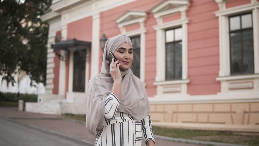 Arab Muslim woman in hijab with makeup walking on the street talking on the phone on a background of beautiful old classic building. The woman is dressed in a stylish clothes