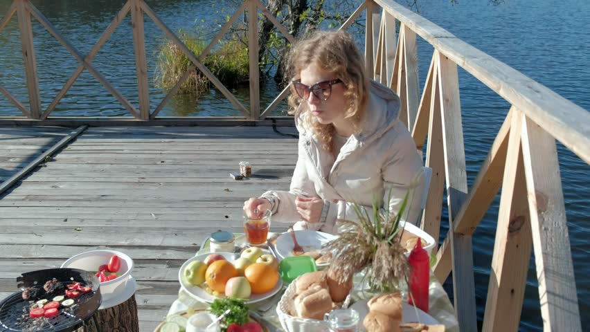 young woman in warm clothes, sitting at the table, drinking tea, eating pancakes with jam, picnic by the river on a wooden bridge, weekend, cold weather, camping, tourism 4k