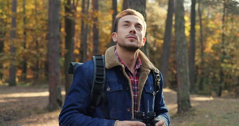 Portrait shot of the Caucasian man standing and looking in the binoculars in the autumn forest, then smiling to the camera. Outdoors. Close up.
