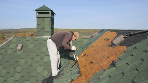Dismantling the soft roof. French green tile. Roofer working on a sloping roof. A man with a beard tears off an old roofing material from a wooden slab with the help of a crowbar. Construction work at