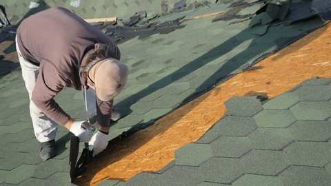 Dismantling the soft roof. French green tile. Roofer working on a sloping roof. A man with a beard tears off an old roofing material from a wooden slab with the help of a crowbar. 