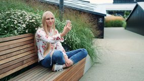 Woman making video call on green roof. Lovely young female using smartphone to make video call while sitting on bench on living roof of huge mall in USA. 4K