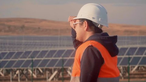 Asian man in an engineer suit on the background of a solar power plant looking at the horizon.
