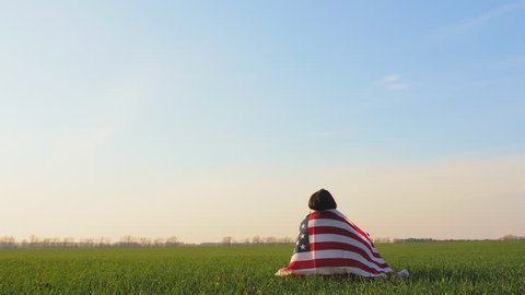 American woman sitting on green grass in a field wrapped a large United States flag