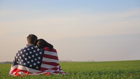 Patriotic American couple wrapped by the flag of America sit on green grass at sunrise in the field