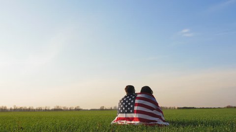 Patriotic man and woman wrapped by the flag of America sit on green grass at sunrise