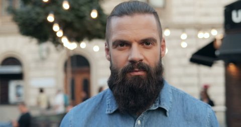 Portrait of the handsome Caucasian young man with long beard and in jeans blue shirt standing on the street of a town with lights and looking with shrewd eyes straight to the camera. Close up. Outdoor