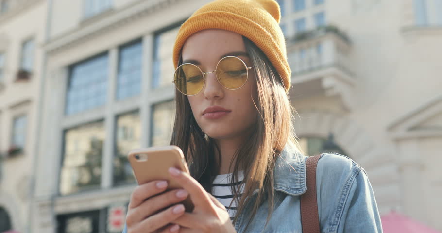 Portrait of the young stylish pretty woman in a cat and sunglasses texting while chatting on the smartphone and smiling on the street. Close up. Outside. | Shutterstock HD Video #1018904917