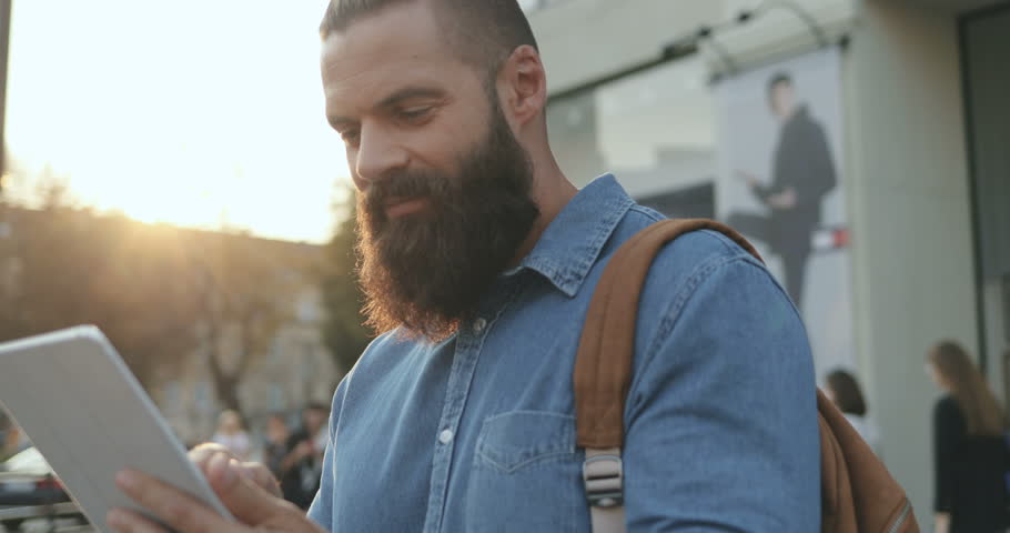 Portrait shot of the good looking young Caucasian man with a beard and backpack using tablet device while standing on the city street. Oudoors.