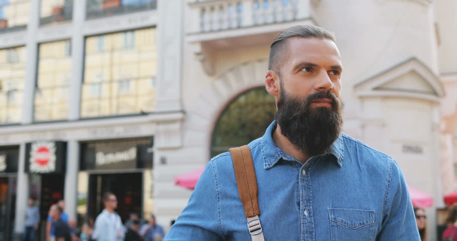 Portrait shot of the handsome Caucasian hipster man with a beard and backpack standing in the city center, looking at the side and then smiling to the camera. Outdoors.