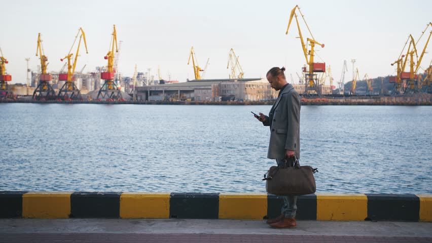 Portrait of young handsome hipster man with bag standing using his mobile phone with sea port on background