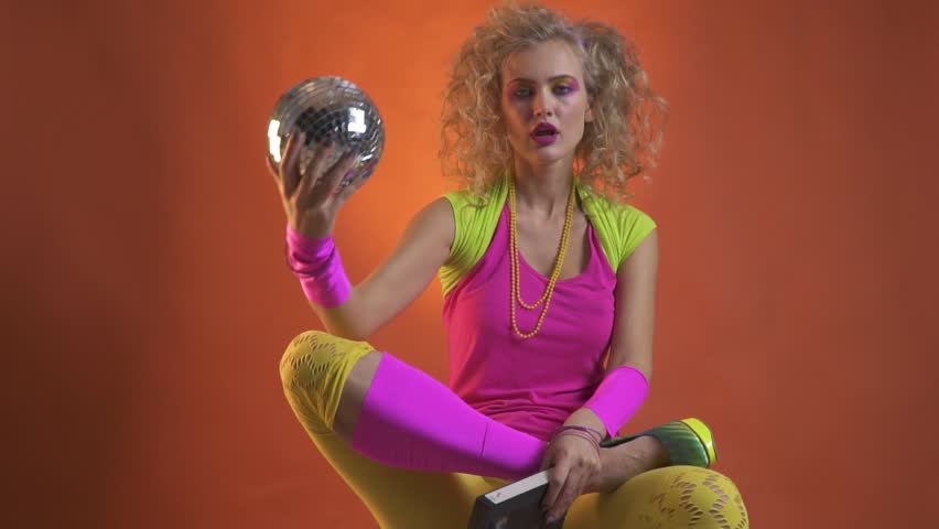 Young woman dressed in retro style clothes, holding a disco ball and a cassette in her hands while dancing with her shoulders