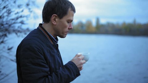 Pensive Man Drinks Green Tea Standing on the River Bank on an Autumn Evening