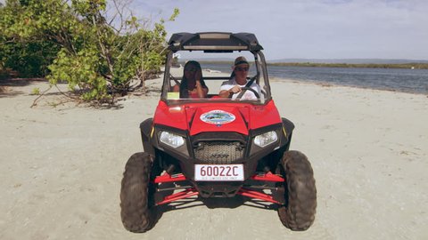 Couple in ATV vehicle driving on beach and under trees with lake in the background in Australia. Medium close on 4k RED camera. Adlı Stok Video