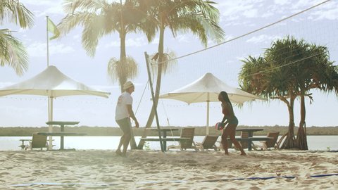 Young couple on vacation playing beach volleyball with ocean, palm trees and in umbrellas in the background in Australia. Wide shot on 4k RED camera.