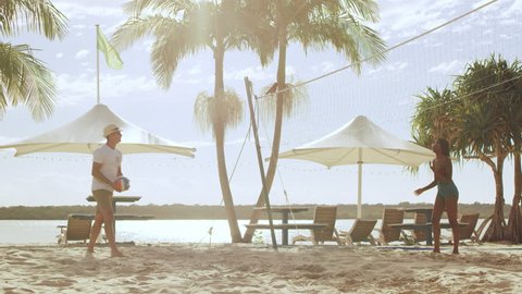 Young couple on vacation playing beach volleyball with ocean, palm trees and in umbrellas in the background in Australia. Wide shot on 4k RED camera. Stockvideo
