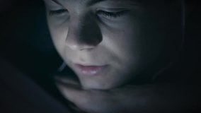 Boy watching movie or playing games on tablet computer at night. Child with headphones under blanket on bed using smartphone or tablet pc. Caucasian boy to make video call to talk to friends. 