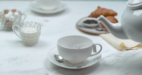 Hot water being poured in a white tea cup, on a fancy table, with croissants, sugar and milk in the background. Medium shot in slow motion with a shallow depth of field in 4K on a Phantom Flex camera.