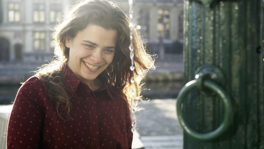 Pretty girl splashing in fountain on central square of Ghent. Portrait playful and smile woman have fun with water in morning sun. Backlight up to illuminate hands and flying in slowmotion drops