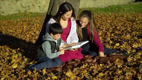 Mom and children are sitting under a tree and reading a book. Mom and children in the autumn forest. They are happy.
