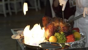 cook is cutting the baked chicken on the plate with vegetables near the fire on the summer terrace of the restaurant