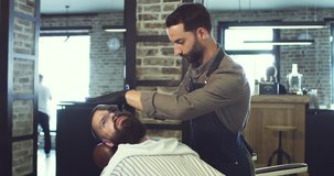 Caucasian profesional barber shaving a beard of his client in the barber shop during his workday.