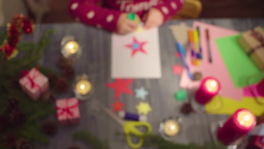 Crane shot. Top view. Little girl glues New Year card. Clip out of focus at the beggining, then focused on the postcard. French text on a sweater - falls