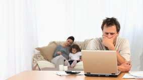 Angry man on his laptop while his family is on a sofa