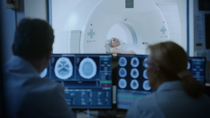 In Control Room Doctor and Radiologist Discuss Diagnosis while Watching Procedure and Monitors Showing Brain Scans Results, In the Background Patient Undergoes MRI or CT Scan Procedure. Royalty-Free Stock Footage #1019002291