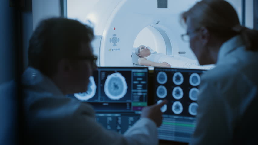 In Control Room Doctor and Radiologist Discuss Diagnosis while Watching Procedure and Monitors Showing Brain Scans Results, In the Background Patient Undergoes MRI or CT Scan Procedure. Royalty-Free Stock Footage #1019002300