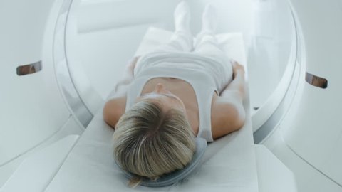 Female Patient Lying on a CT or MRI Scan, Bed is Moving inside Machine Scanning Her Body and Brain. In Medical Laboratory with High-Tech Equipment. Elevated Camera Shot.