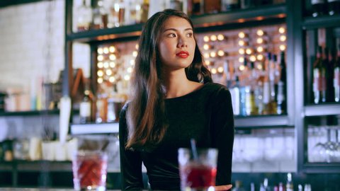Portrait of an attractive Asian woman smiling and standing by a bar with drinks in interior cool bar with soft interior lighting. Medium shot on 4k RED camera.