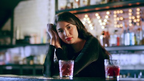 Portrait of an attractive Asian woman leaning on a bar with drinks in interior cool bar with soft interior lighting. Medium shot on 4k RED camera.