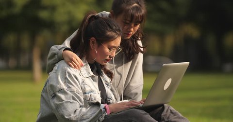 Two happy asian college girl sitting on the lawn using laptop and talking together in campus.
