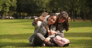 Slow motion of two asian teen girl sitting on the lawn using mobile phone taking selfie , video chatting outdoor in the park
