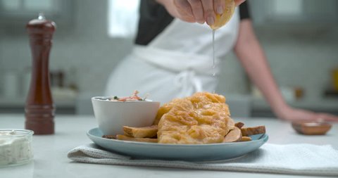 A chef squeezes a half cut lemon over a plate of gourmet battered fish and chips in interior restaurant kitchen in soft light. Medium shot at 1000 fps in 4k on a Phantom Flex camera
