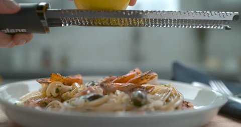 A chef's hand grates a lemon over a white plate of shrimp and olive linguini in interior restaurant kitchen with soft light. Closeup in 4k at 1000 fps on a Phantom Flex camera