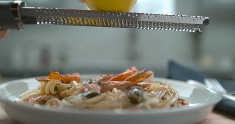 A chef's hand grates a lemon over a white plate of shrimp and olive linguini in interior restaurant kitchen with soft light. Closeup in 4k at 1000 fps on a Phantom Flex camera