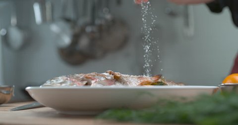 A chef's hand sprinkles rock salt onto a plate of fish, on a wooden chopping board with herbs in a restaurant kitchen in slow motion, in soft light. Closeup in 4k at 1000 fps on a Phantom Flex camera