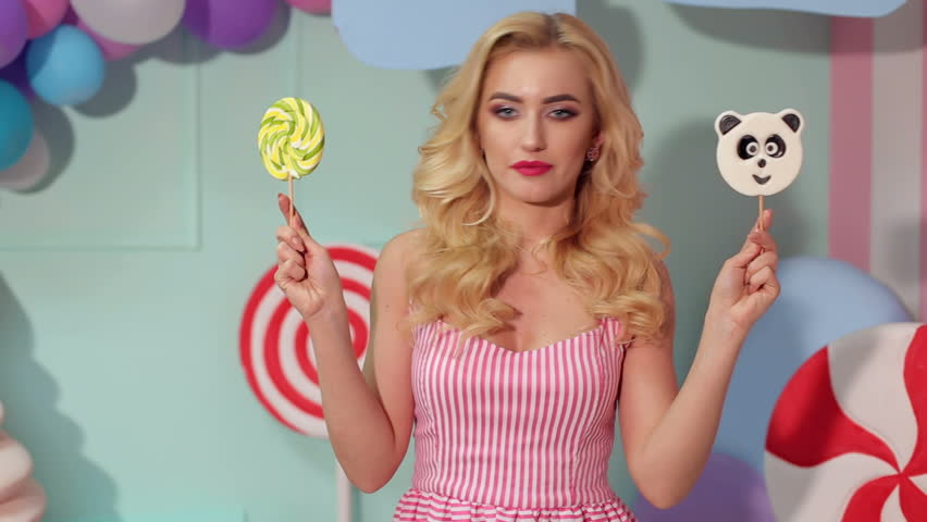 A cheerful girl with big lollipops in her hands in a bright room with balloons and a large plastic ice cream, she covers her eyes with big lollipops. Barbie girl. Candy girl.