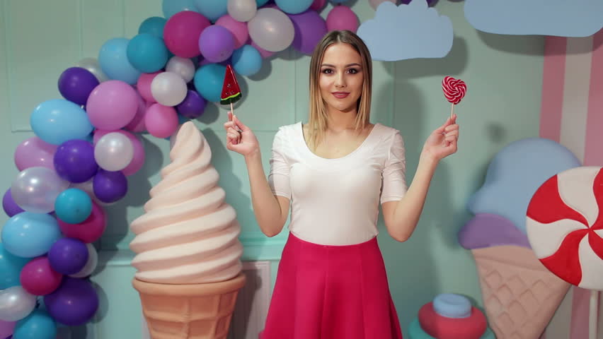 A young laughing girl holds a big candy in her hands, she covers her eyes with big lollipops on the background of colorful balloons in a bright studio.