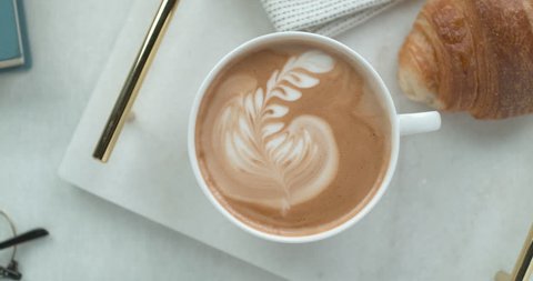 Pretty latte leaf design in white cup next to butter croissant and reading glasses ultra slow motion closeup