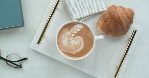 Pretty latte leaf design in white cup next to butter croissant and reading glasses ultra slow motion